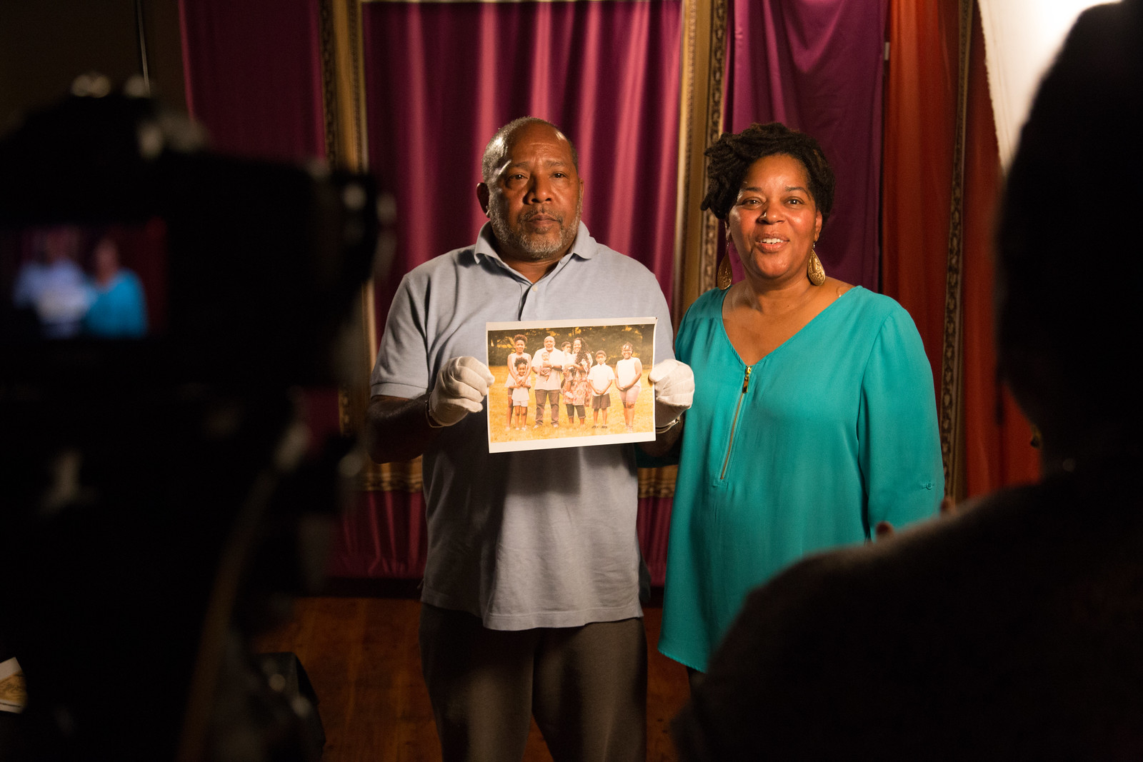 A man and woman posing in front of a camera, holding a family photograph